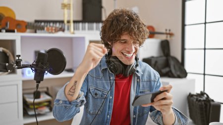 Photo for A jubilant young man with headphones in a music studio celebrates good news on his smartphone. - Royalty Free Image