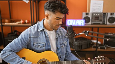 Handsome hispanic man playing guitar in a music studio, microphone in foreground, 'on air' sign illuminated in background.