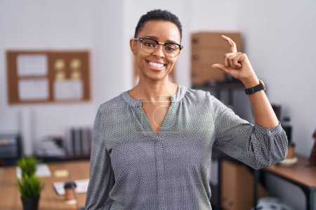 Téléchargez les photos : Femme afro-américaine travaillant au bureau portant des lunettes geste souriant et confiant avec la main faisant signe de petite taille avec les doigts regardant et la caméra. concept de mesure. - en image libre de droit