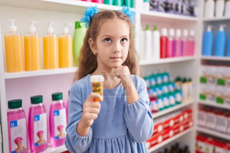Photo for Young little girl holding pills at the pharmacy serious face thinking about question with hand on chin, thoughtful about confusing idea - Royalty Free Image