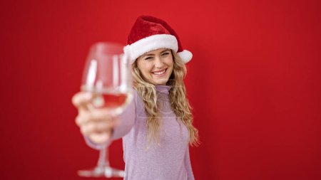Foto de Mujer rubia joven con sombrero de Navidad sosteniendo una copa de vino sobre un fondo rojo aislado - Imagen libre de derechos