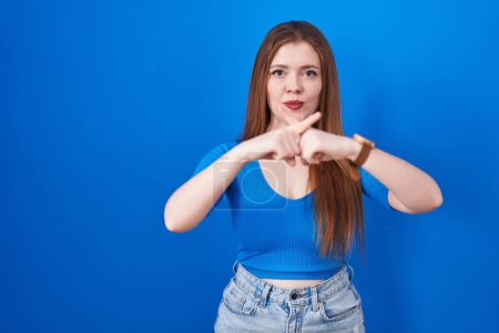 Photo for Redhead woman standing over blue background rejection expression crossing fingers doing negative sign - Royalty Free Image