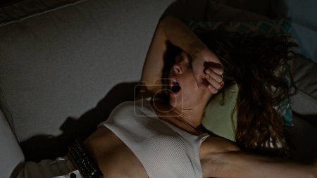 Téléchargez les photos : Une jeune femme fatiguée bâillant alors qu'elle était allongée sur un canapé avec une télécommande à la main la nuit dans une pièce faiblement éclairée. - en image libre de droit