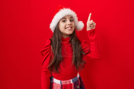 Téléchargez les photos : Adorable fille hispanique dans le chapeau de Noël, pointant joyeusement le doigt vers le haut, signalant le succès de l'idée numéro un sur fond rouge vibrant isolé - en image libre de droit