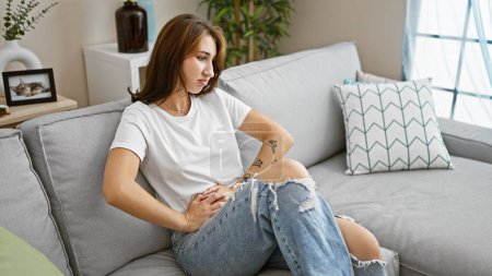 Téléchargez les photos : Jeune femme souffrant de maux d'estomac assis sur le canapé à la maison - en image libre de droit