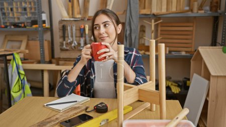 Photo for Hispanic woman enjoying a coffee break in a woodworking workshop, surrounded by tools and wooden furniture. - Royalty Free Image