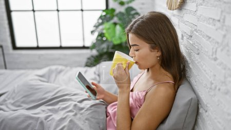 Téléchargez les photos : Jolie jeune femme hispanique assise détendue en pyjama sur un lit confortable, sirotant sereinement le café du matin tout en envoyant des SMS sur smartphone dans sa chambre confortable. - en image libre de droit