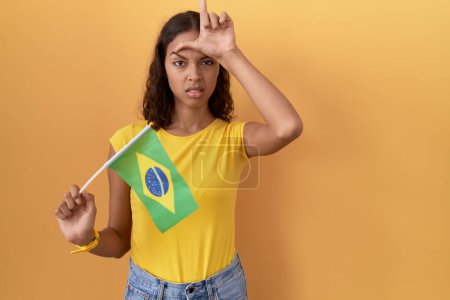 Photo for Young hispanic woman holding brazil flag making fun of people with fingers on forehead doing loser gesture mocking and insulting. - Royalty Free Image