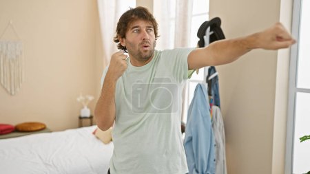 Téléchargez les photos : Homme barbu dans un t-shirt décontracté imite un coup de poing dans une chambre moderne. - en image libre de droit