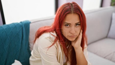 Photo for Worried redhead adult sitting on the home sofa caught in a web of doubt, a sad expression painting the beautiful young woman's portrait in her living room - Royalty Free Image