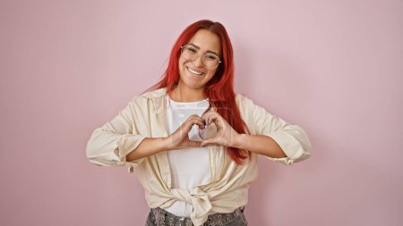 Photo for Cheerful young redhead woman, smiling confidently & gesturing a love heart with hands over isolated pink background - Royalty Free Image