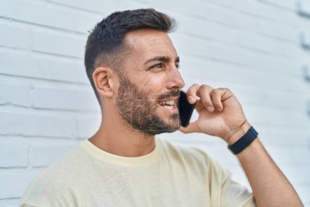 Téléchargez les photos : Jeune homme hispanique souriant confiant parler sur le smartphone sur fond blanc isolé - en image libre de droit