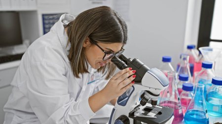 Foto de Una científica hispana enfocada examina muestras bajo un microscopio en un laboratorio bien equipado. - Imagen libre de derechos