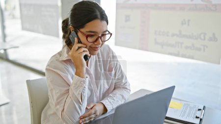 Foto de Mujer hispana conversa por teléfono en el entorno moderno de la oficina, portátil y portapapeles en el escritorio. - Imagen libre de derechos