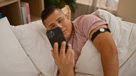 Foto de Young latin man using smartphone lying on bed at bedroom - Imagen libre de derechos