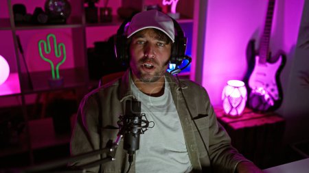 Foto de Hombre guapo con auriculares en una colorida sala de juegos iluminada por neón por la noche, hablando en un micrófono. - Imagen libre de derechos