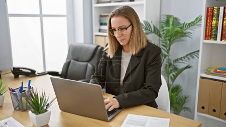 Téléchargez les photos : Jeune femme d'affaires blonde dédiée travaillant intensément sur son ordinateur portable dans un cadre de bureau professionnel - en image libre de droit