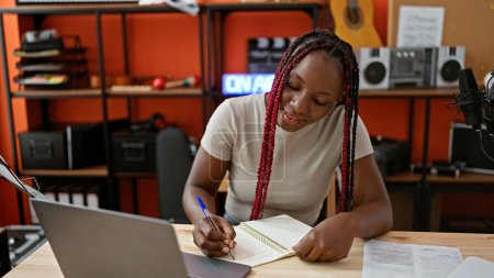 Téléchargez les photos : Souriante musicienne afro-américaine prenant magistralement des notes sur ordinateur portable lors d'un appel vidéo passionnant dans un studio de musique dynamique - en image libre de droit