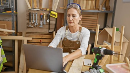 Photo for A mature woman in a woodworking studio focuses intently on her laptop amidst various carpentry tools. - Royalty Free Image