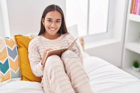 Téléchargez les photos : Young beautiful hispanic woman writing on notebook sitting on bed at bedroom - en image libre de droit
