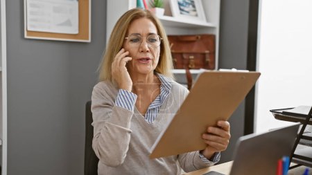 Téléchargez les photos : Une femme professionnelle discute au téléphone tout en examinant un document dans son bureau moderne. - en image libre de droit