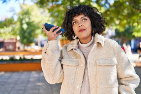 Photo for Young beautiful hispanic woman listening audio message by the smartphone with serious expression at park - Royalty Free Image