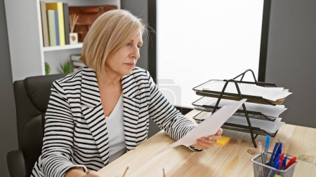 Téléchargez les photos : Femme blonde mature examinant attentivement un document dans un cadre de bureau moderne. - en image libre de droit