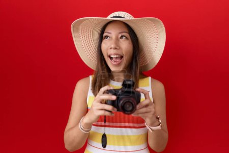 Foto de Mujer china de mediana edad usando cámara réflex usando sombrero de verano enojado y loco gritando frustrado y furioso, gritando con ira mirando hacia arriba. - Imagen libre de derechos