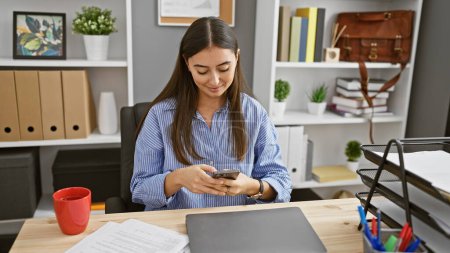 Photo for Hispanic woman texting in a modern office, surrounded by shelves, plants, and desk accessories. - Royalty Free Image