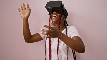 Photo for Jolly african american woman gaming away, standing confidently over pink isolated background, playing video game with virtual reality glasses - Royalty Free Image