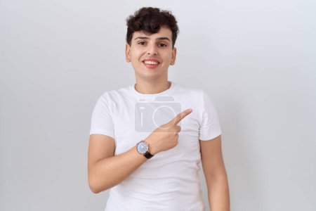 Téléchargez les photos : Jeune homme non binaire portant un t-shirt blanc décontracté gai avec un sourire sur le visage pointant avec la main et le doigt sur le côté avec une expression heureuse et naturelle - en image libre de droit