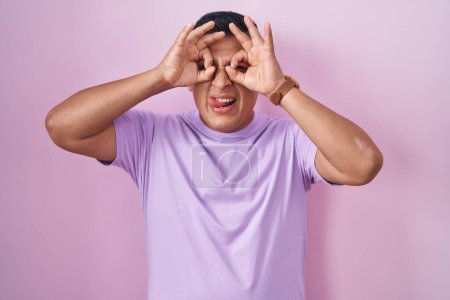 Foto de Un joven hispano parado sobre un fondo rosado haciendo un buen gesto como binoculares sacando la lengua, ojos mirando a través de los dedos. expresión loca. - Imagen libre de derechos