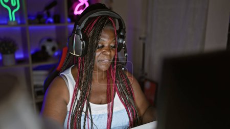 Foto de Confiada mujer afroamericana streamer jugando alegremente a videojuegos en su acogedora sala de juegos, inmersa en el entretenimiento cibernético futurista por la noche - Imagen libre de derechos
