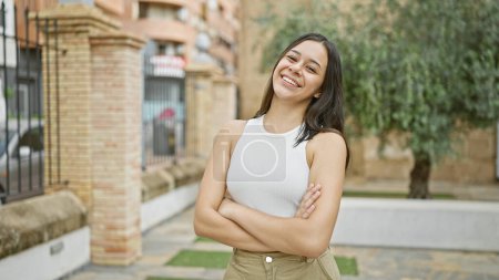 Foto de Joven hermosa mujer hispana sonriendo confiada de pie con los brazos cruzados gesto en el parque - Imagen libre de derechos