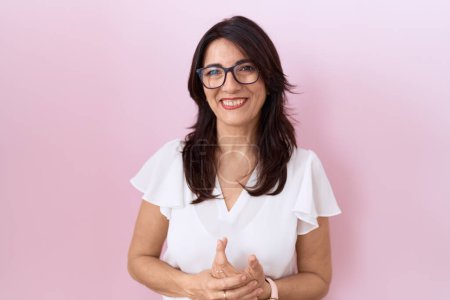Téléchargez les photos : Femme hispanique du Moyen Âge portant un t-shirt blanc décontracté et des lunettes avec les mains jointes et les doigts croisés souriant détendu et joyeux. succès et optimisme - en image libre de droit