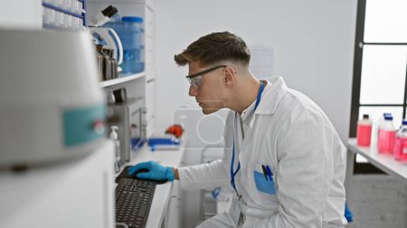 Photo for Attractive young caucasian male scientist totally engrossed in typing on computer amidst test tubes and microscopes, indoor laboratory work - Royalty Free Image