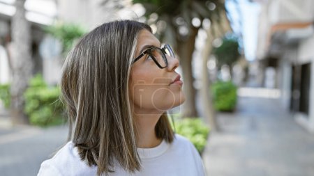 Téléchargez les photos : Une jeune femme hispanique avec des cheveux bruns et des lunettes regarde attentivement tout en se tenant debout dans une rue de la ville. - en image libre de droit