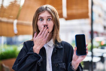 Photo for Shocked blonde beauty covers mouth in surprise, young woman's fearful expression caught on cellphone at urban coffee shop terrace - Royalty Free Image