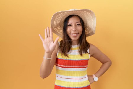 Foto de Mujer china de mediana edad con sombrero de verano sobre fondo amarillo renunciando a decir hola feliz y sonriente, gesto de bienvenida amistoso - Imagen libre de derechos