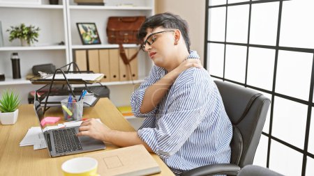 Foto de Hombre que experimenta dolor de cuello mientras trabaja en un ordenador portátil en un entorno de oficina moderno, que representa el malestar en el lugar de trabajo. - Imagen libre de derechos