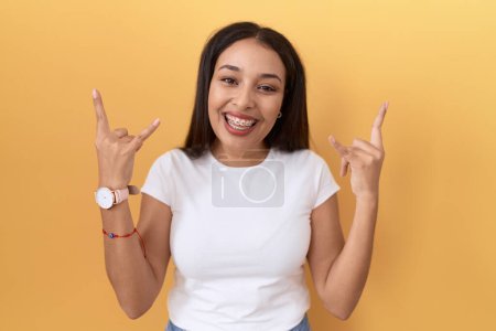 Foto de Mujer árabe joven con camiseta blanca casual sobre fondo amarillo gritando con expresión loca haciendo símbolo de roca con las manos arriba. Estrella de la música. concepto de música pesada. - Imagen libre de derechos