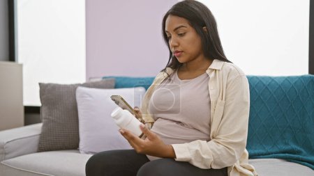 Foto de Mujer hispana joven preocupada, embarazada y enferma, pegada a su teléfono inteligente en casa, consultando nerviosamente la información de la píldora - Imagen libre de derechos
