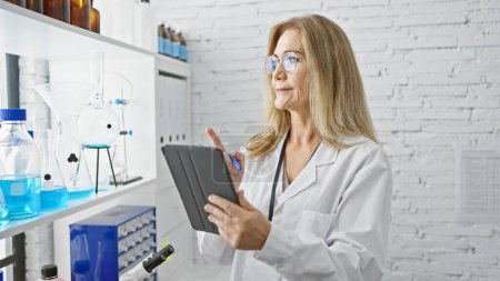 Foto de Una científica madura analiza los datos de una tableta en un laboratorio blanco con varios productos químicos y equipos. - Imagen libre de derechos