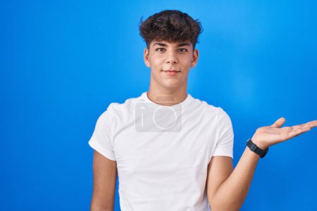 Téléchargez les photos : Adolescent hispanique debout sur fond bleu souriant présentation joyeuse et pointant avec la paume de la main en regardant la caméra. - en image libre de droit