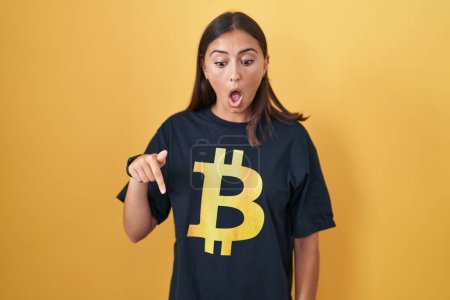 Photo for Young hispanic woman wearing bitcoin t shirt pointing down with fingers showing advertisement, surprised face and open mouth - Royalty Free Image