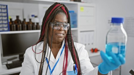 Foto de Confiada científica afroamericana con trenzas sonriendo mientras sostiene una botella llena de líquido en un laboratorio médico, abrazando la belleza de la ciencia y la tecnología - Imagen libre de derechos