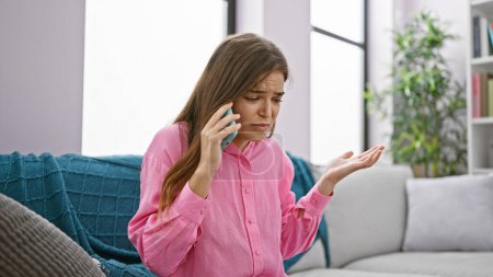 Photo for Unhappy, worried young beautiful hispanic woman talking with serious problems via smartphone, sitting alone at home on the living room sofa. - Royalty Free Image