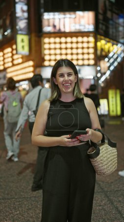 Photo for Beautiful hispanic woman smiling at tokyo's urban night lights while texting happily on her smartphone - Royalty Free Image