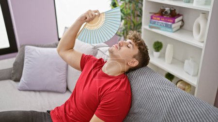 Téléchargez les photos : Malheurs du temps chaud, jeune homme hispanique souffre, se ventilant avec un ventilateur assis sur le canapé à la maison, exposant les luttes de la chaleur intérieure - en image libre de droit