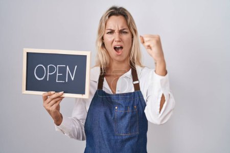 Photo for Young blonde woman wearing apron holding blackboard with open word annoyed and frustrated shouting with anger, yelling crazy with anger and hand raised - Royalty Free Image